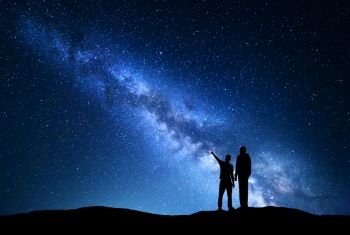 Milky Way with silhouette of men. Father and a son who pointing finger in night starry sky on the mountain. Night landscape. Silhouette of family on the background of beautiful galaxy. Space. 
