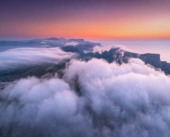 Aerial view of low clouds, mountains, sea and colorful sky at sunset. Above the clouds at dusk. Seashore in Spain. Top view from drone. Amazing landscape with cloudy sky and rocks. Nature background