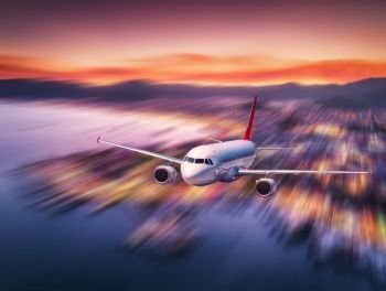 Airplane with motion blur effect is flying over sea coast at night. Landscape with passenger airplane, blurred buildings, city illumination, sea and sky. Aircraft. Business travel. Commercial plane