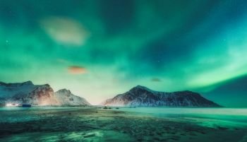 Northern lights above the rocks in Lofoten islands, Norway. Aurora borealis. Starry sky with polar lights and clouds. Night winter landscape with aurora, sea, beach and snowy mountains. Travel