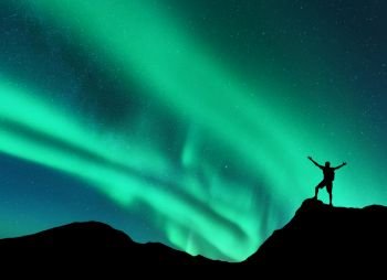 Northern lights and silhouette of standing man with raised up arms on the mountain peak in Norway. Aurora borealis and happy man. Sky with stars and green polar lights. Night landscape with aurora