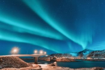 Northern lights above bridge with illumination in Lofoten islands, Norway. Aurora borealis. Starry sky with polar lights. Night winter landscape with aurora, road, village and snowy mountains. Travel