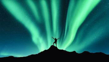 Northern lights and silhouette of standing man with raised up arms on the mountain in Norway. Aurora borealis and happy man. Sky with stars and green polar lights. Night landscape with aurora. Concept