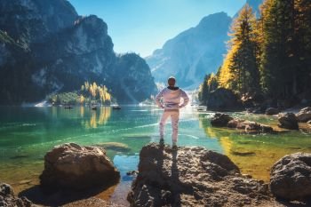 Standing sporty man on the stone on the coast of Braies lake at sunrise. Autumn in Dolomites, Italy. Landscape with happy man, mountains, water with reflection, fall trees and blue sky in the morning