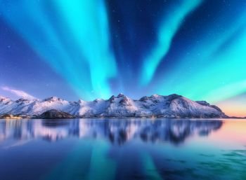 Northern lights and snow covered mountains in Lofoten islands, Norway. Aurora borealis. Starry sky with polar lights and snowy rocks reflected in water. Night winter landscape with aurora, sea. Travel. Northern lights and snow covered mountains in Lofoten islands, Norway