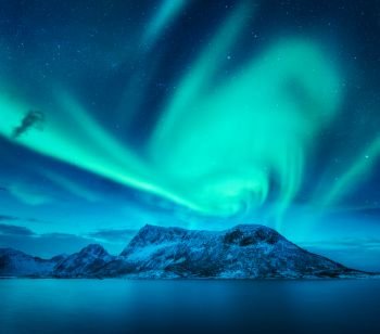 Aurora borealis above the snow covered mountain in Lofoten islands, Norway. Northern lights in winter. Night landscape with green polar lights, snowy rocks, blue sea. Beautiful starry sky with aurora. Aurora borealis above the snow covered mountain