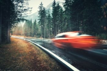 Blurred red car in motion on the road in autumn forest in rain. Perfect asphalt mountain road in overcast rainy day. Roadway, pine trees in italian alps. Transportation. Highway in foggy woodland
