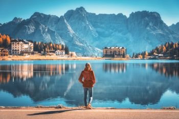 Young woman standing on the coast of Misurina lake at sunrise in autumn. Dolomites, Italy. Landscape with girl in red jacket, reflection in water, buildings, blue sky. Italian alps. Travel background
