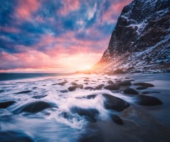 Sandy beach with stones in blurred water, blue sky with pink clouds and snowy mountains at sunset. Utakleiv beach, Lofoten islands, Norway. Winter landscape with sea, waves, rocks in the evening