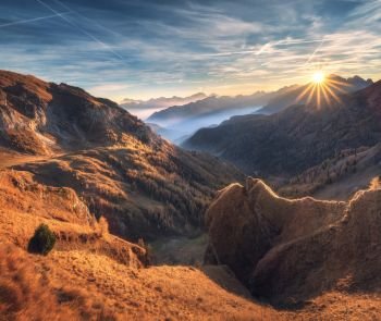 Mountains in fog at beautiful sunset in autumn. Dolomites, Italy. Landscape with alpine mountain valley, orange grass, low clouds, trees on hills, blue sky with clouds and sun. Aerial view. Passo Giau