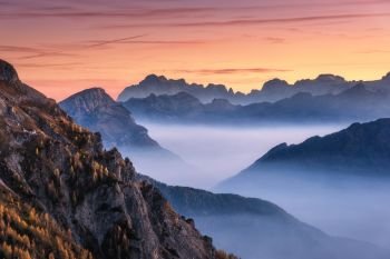 Mountains in fog at beautiful sunset in autumn in Dolomites, Italy. Landscape with alpine mountain valley, low clouds, trees on hills, orange sky with clouds at dusk. Aerial view. Passo Giau. Nature