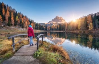 Young wooman on the small wooden bridge against lake, autumn forest, mountain peak, blue sky with sunlight at sunrise. Landscape with girl, reflection in water, green grass, rocks in Dolomites, Italy
