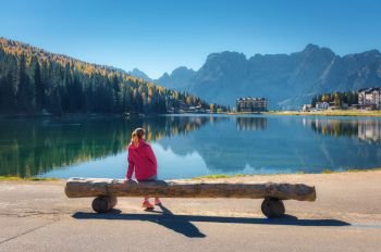 Young woman sitting on the wooden bench on the coast of Misurina lake at sunset in autumn. Dolomites, Italy. Landscape with girl in red jacket, reflection in water, forest, buildings, blue sky. 