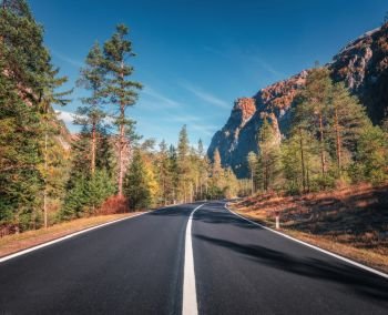 Road in autumn forest at sunset in Italy. Beautiful mountain roadway, green tress, orange grass, high rocks, blue sky. Landscape with empty asphalt road through the forest in fall. Travel background