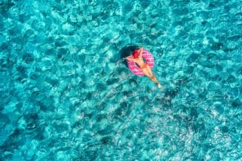 Aerial view of slim young woman swimming on the pink donut swim ring in the transparent blue sea at sunny bright day. Tropical aerial landscape with girl, azure water. Top view. Summer travel. Holiday