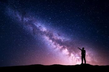Milky Way with silhouette of a standing young man pointing finger in night starry sky on the mountain. Night landscape. Beautiful Universe, travel background with purple sky full of stars and yellow light