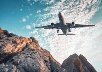 Big airplane is flying over rocks at sunset in summer. Landscape with  landing passenger airplane, mountains, colorful blue sky with clouds. Business travel. Commercial plane. Vintage toning. Aircraft