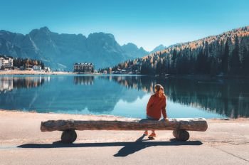 Young woman sitting on the wooden bench on the coast of Misurina lake at sunset in autumn. Dolomites, Italy. Landscape with girl in red jacket, reflection in water, forest, buildings, blue sky. 