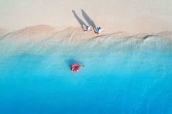 Aerial view of a young woman swimming with the donut swim ring in the blue sea, waves and walking people at sunset in summer. Tropical aerial landscape with girl, azure water, sandy beach. Top view