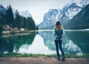 Young woman with backpack is standing on the coast of mountain lake at cloudy day in spring. Travel in Italy. Landscape with slim girl, reflection in water, snowy rocks, green trees. Vintage toning. Young woman with backpack is standing on the coast of the lake 