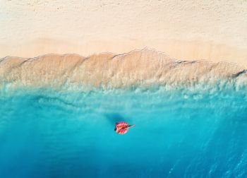 Aerial view of a young woman swimming with the donut swim ring in the clear blue sea with waves at sunset in summer. Tropical aerial landscape with girl, azure water, sandy beach. Top view. Travel. Aerial view of a swimming woman in the sea at sunset