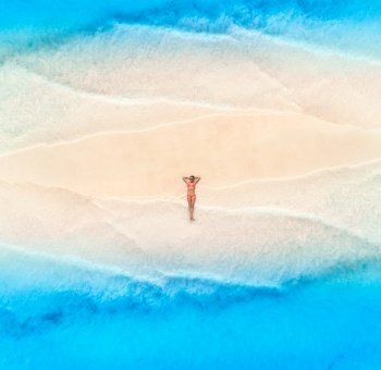 Aerial view of the beautiful young lying woman on the white sandy beach and blue sea with waves on the both sides at sunset. Summer holiday. Top view of sporty slim girl, sandbank, clear water