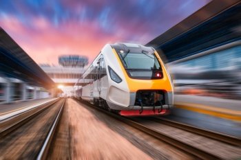 High speed train in motion on the railway station at sunset. Fast moving modern passenger train on railway platform. Railroad with motion blur effect. Commercial transportation. Front view. Concept
