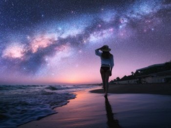 Milky Way and young woman in hat on sandy beach against purple starry sky reflected in water at night. Landscape. Summer travel. Girl on the tropical sea coast and bright Milky Way and stars. Space	