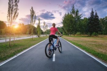 Woman riding a mountain bike on the asphalt road at sunset in summer. Colorful landscape with sporty girl riding a bicycle, road, green trees, grass and gold sunlite in park. Sport and travel. Cycle
