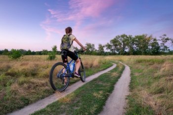 Woman is riding a mountain bike in cross country road at sunset in summer. Colorful landscape with sporty girl with backpack riding bicycle, field, dirt road, green grass, purple sky. Sport and travel