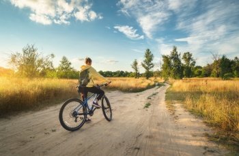 Woman is riding a mountain bike in cross country road at sunset in summer. Colorful landscape with sporty girl with backpack riding bicycle, field, dirt road, green grass, blue sky. Sport and travel