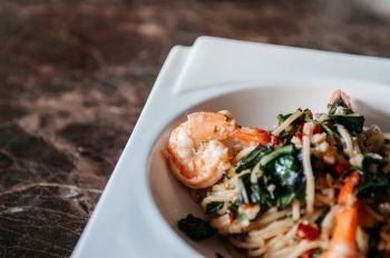 Fusion Pasta spicy Thai Pad Kapraw spaghetti with garlic, red chilli, shrimps and holy basil in white plate on marble table.