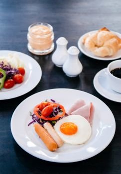 A set of American breakfast sunny side up eggs, sausage, ham, fresh salad bell peppers, tomato, croissant bread with black coffee