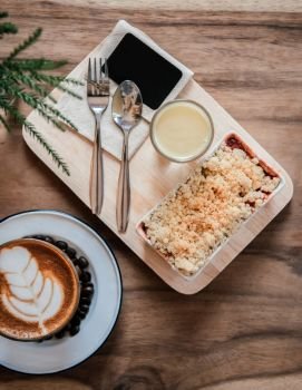 Berry and apple crumble on wood tray with hot latte coffee on wood table. Top view food shot