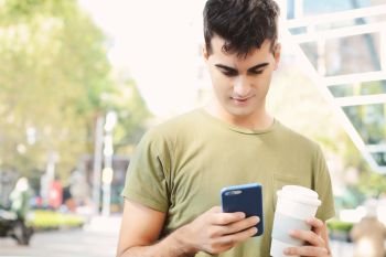 Young man holding coffee and reading message on his phone. Outdoors.