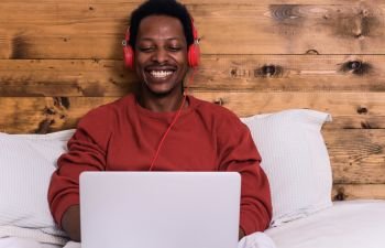 Young afro american man using his laptop with headphones in bed.