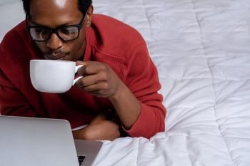 Young afro american man using his laptop with cup of coffee in bed.