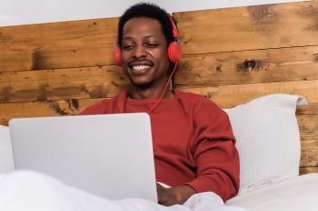 Young afro american man using his laptop with headphones in bed.