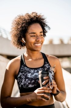 Portrait of afro athlete woman holding a bottle of water and relaxing after work out outdoors. Sport and healthy lifestyle.