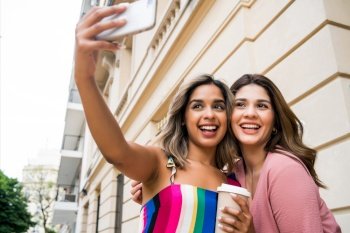 Two young friends smiling and taking a selfie with their mobile phone while standing outdoors. Urban concept.