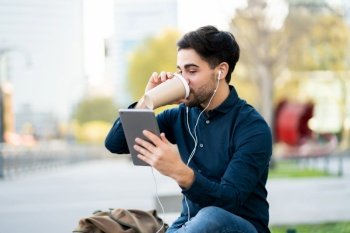 Portrait of young man having a video call on digital tablet and drinking coffee while sitting on bench outdoors. Urban concept.
