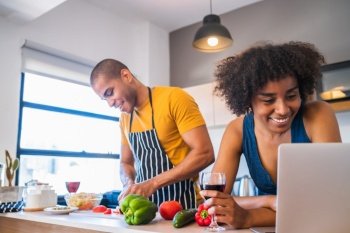 Portrait of young latin couple using a laptop while cooking in kitchen at home. Relationship, cook and lifestyle concept.