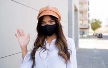 Close-up of young woman wearing protective mask and wave hand to say hello while standing outdoors on the street. Urban concept. New normal lifestyle concept.