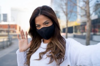 Portrait of young woman wearing face mask and taking selfies while wave hand to say hello outdoors. Urban concept. New normal lifestyle concept.