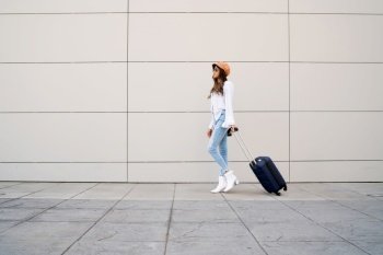 Portrait of young traveler woman carrying a suitcase while walking outdoors on the street. Tourism concept.