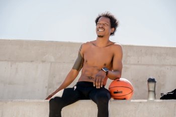Afro athletic man holding a basketball ball and relaxing after training while sitting outdoors. Sport and healthy lifestyle concept.