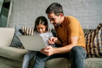 Little girl and her father having fun and using a laptop together while sitting on a couch at home. Monoparental family concept.