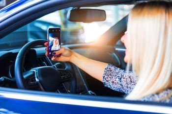 Young happy smiling woman driver taking a selfie photo on a smartphone in her modern luxury car
