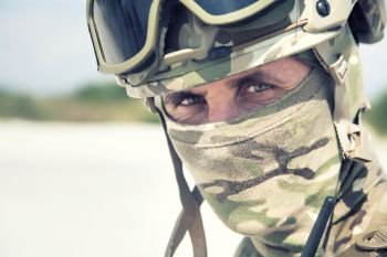 Close up, cropped portrait of airsoft, strikeball war games player, military or historical reenactment participant in tactical helmet replica, with hidden behind camouflage mask face looking at camera. Military softair war games or reenactment participant