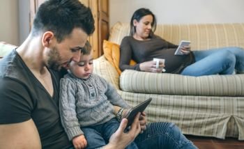 Young father looking at the tablet with his little son while the pregnant mother takes a tea and looks the mobile. Father looking tablet with little son while pregnant mother looks mobile